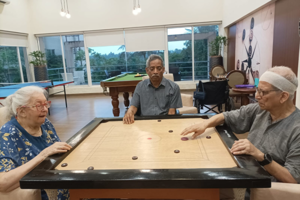 Three senior citizens gathered around a wooden board playing a game of carrom in a bright indoor recreation room.