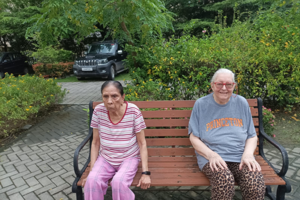 Two elderly women sitting together on a wooden garden bench outdoors surrounded by green plants and trees.