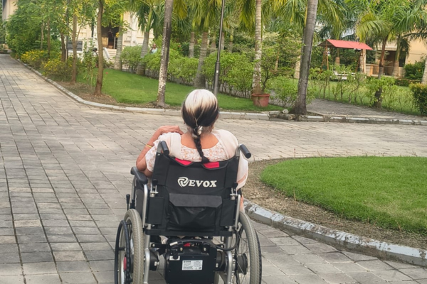 Rear view of an elderly woman with braided hair sitting in an Evox motorized wheelchair on a paved garden path.