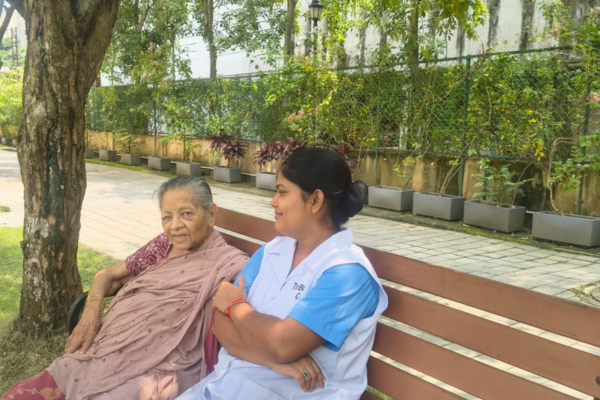 An elderly woman in traditional Indian attire sitting on a wooden park bench alongside a smiling carer in a white uniform.