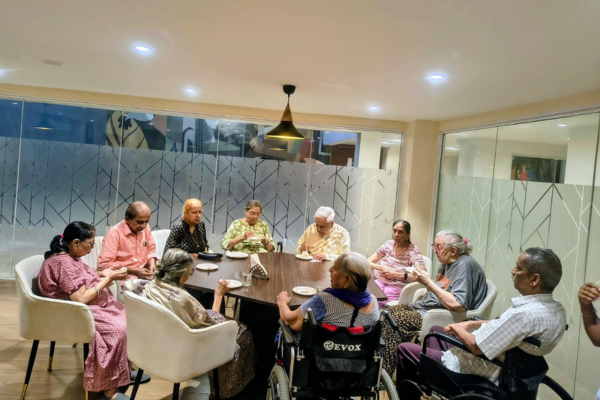 A group of Indian senior citizens sitting around a round table enjoying food together in a well-lit, modern indoor dining room at the Jagriti Dham senior living facility.