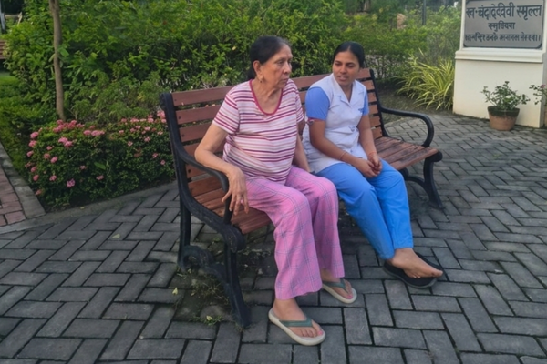 A female caregiver in a white and blue uniform sitting on a wooden garden bench alongside an elderly woman at Jagriti Dham senior living facility.