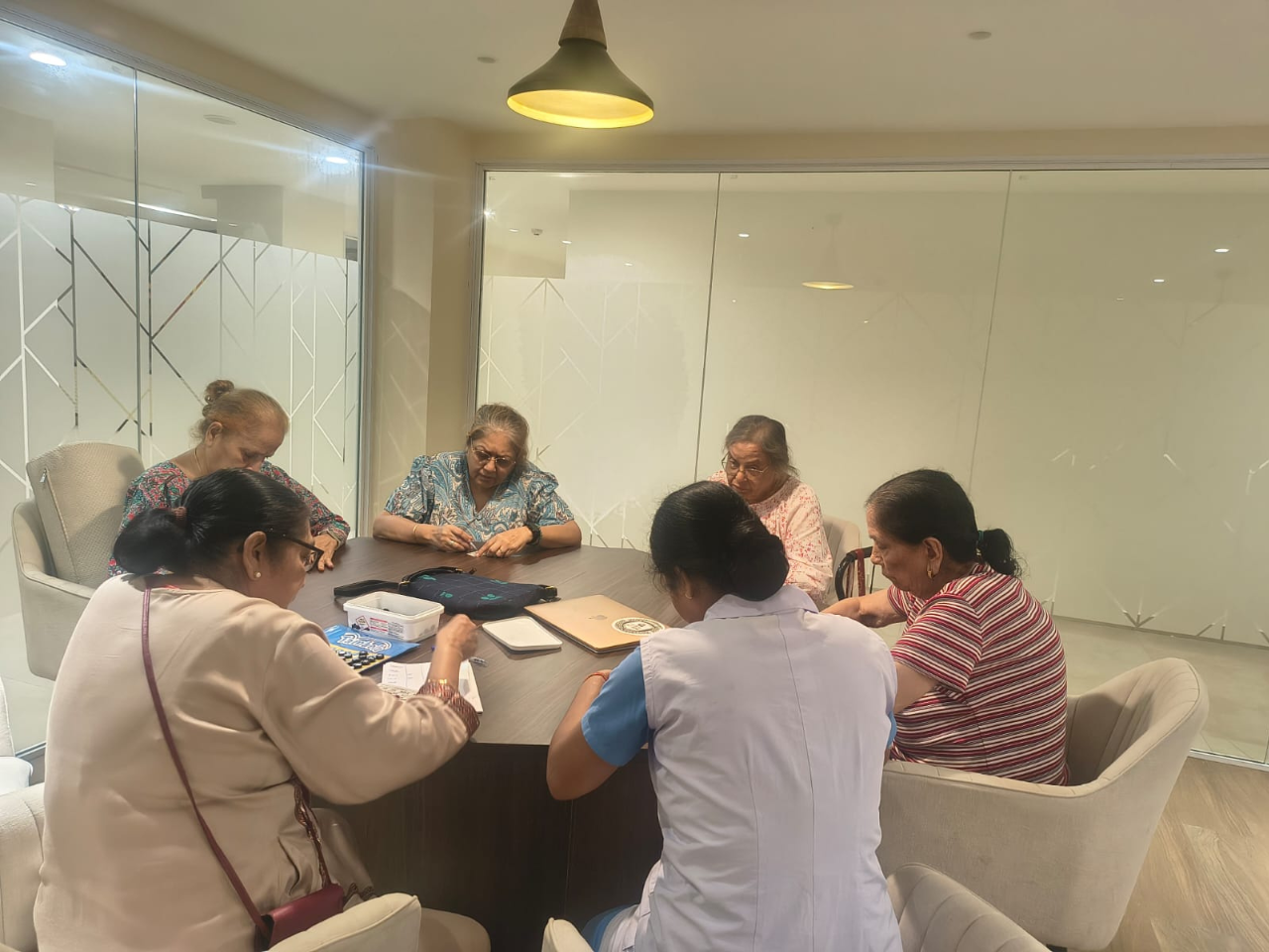 Senior women sitting around a large dark wooden table indoors participating in a group activity with a staff member.