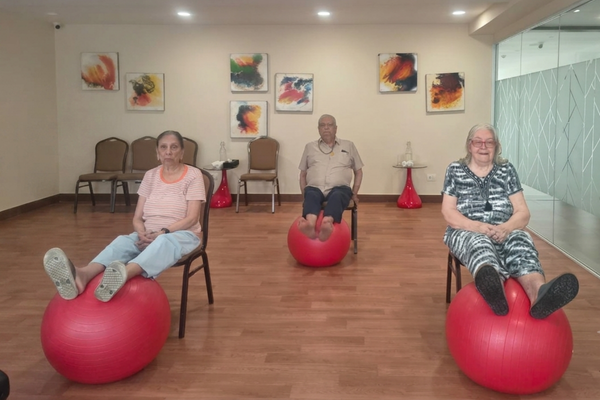 Three seniors sitting on chairs exercising with large red yoga balls under their feet in a modern, well-lit activity room at Jagriti Dham.