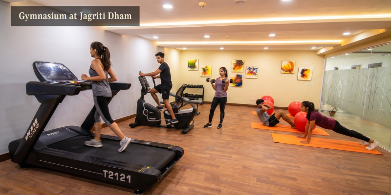 Multiple people engaging in various fitness activities, including treadmill walking, cycling, lifting dumbbells, and stretching, inside a modern gymnasium at Jagriti Dham.