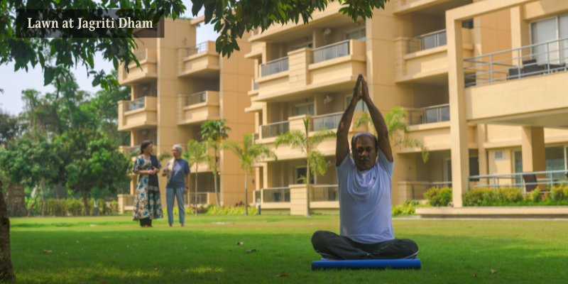 A man performing a seated yoga pose on a mat on a lush green lawn in front of modern residential buildings at Jagriti Dham, with two people conversing in the background.