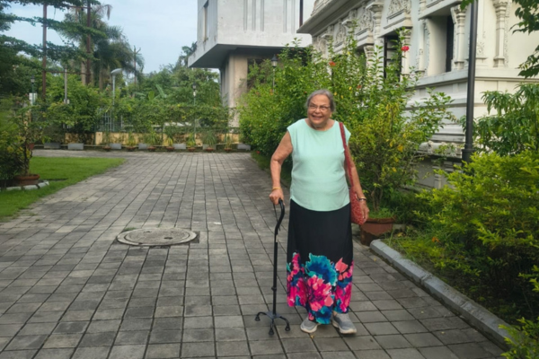 An elderly woman walking happily with a quad cane along a paved outdoor pathway, representing active independent and assisted living.