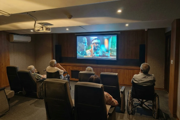 An interior shot of a dark home theatre or screening room where several elderly individuals in armchairs and wheelchairs are watching a movie on a large projector screen.