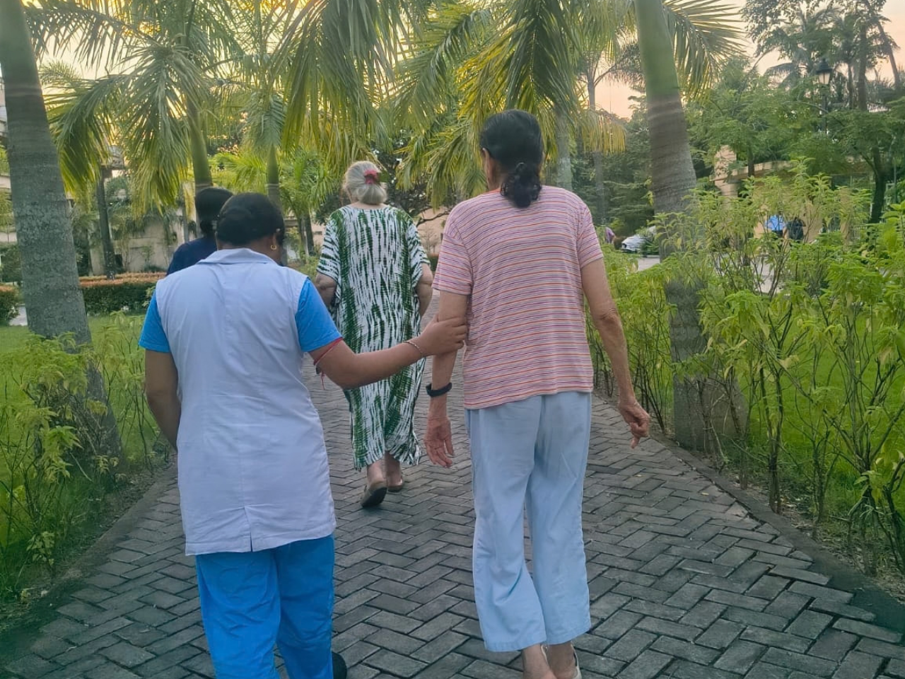 A caregiver in uniform walks with two elderly women in a garden.