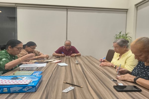 Senior citizens sitting around a wooden table playing a game of Tambola in a brightly lit room.
