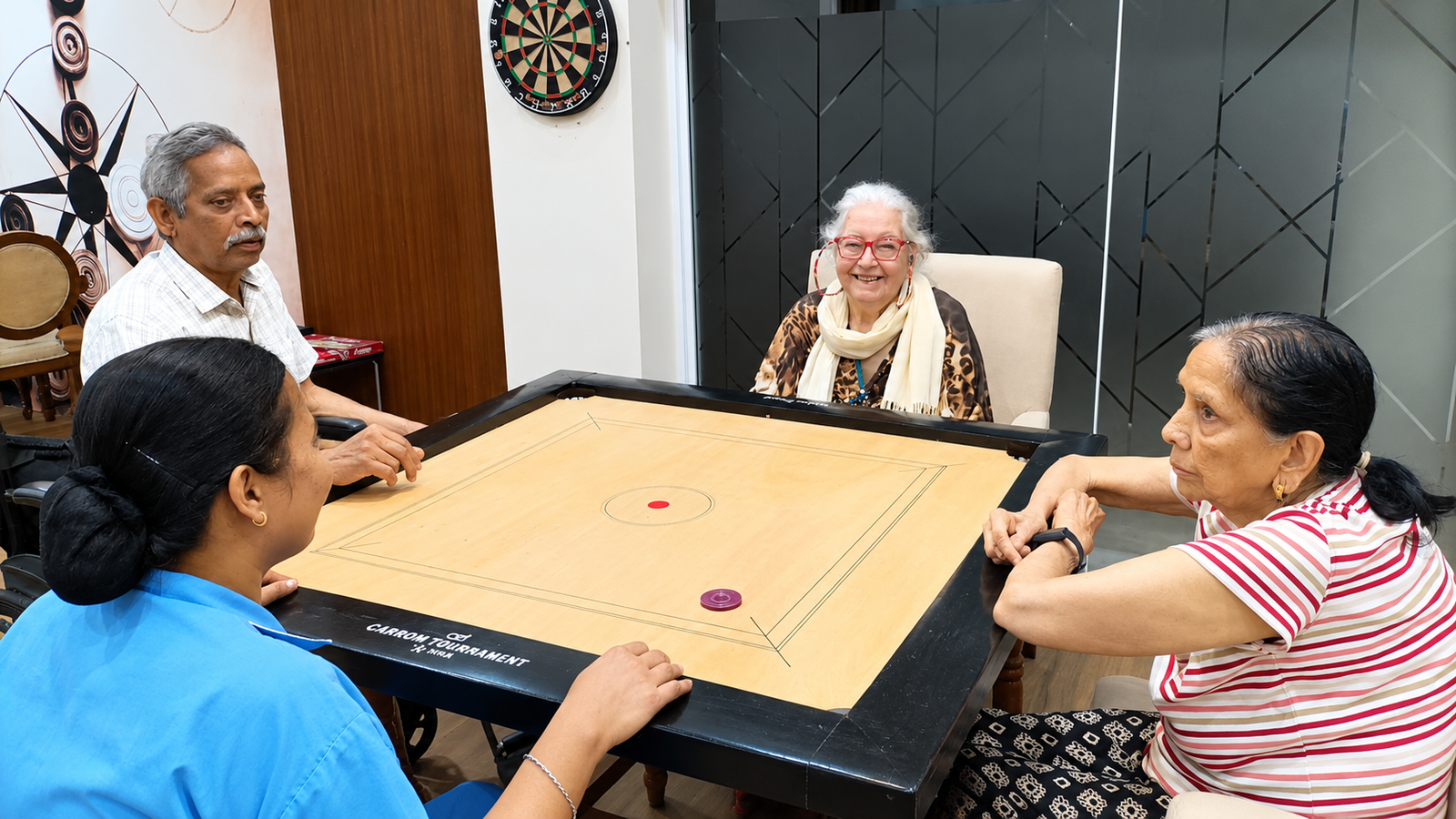 Four older adults enjoying a game of carrom together in the recreation room at Jagriti Dham.