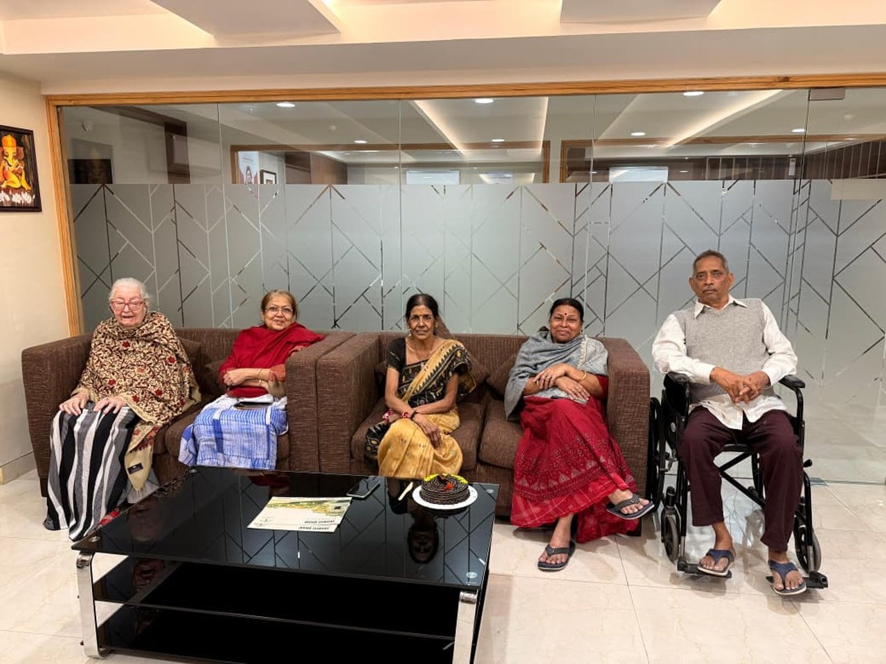 Five senior citizens sitting together in a comfortable lounge with a chocolate cake on the coffee table.